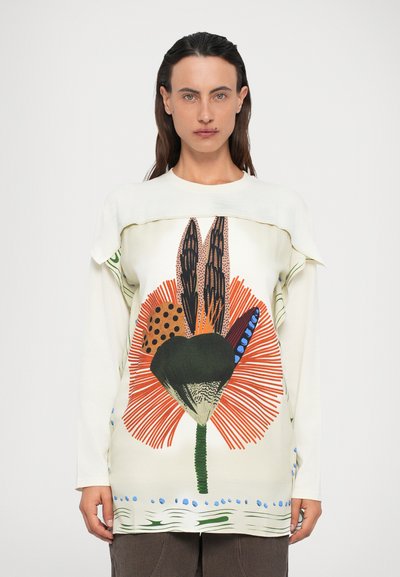 Mujer vestida con una camisa blanca de manga larga con un gran diseño floral abstracto y colorido en el frente, de pie contra un fondo sencillo.