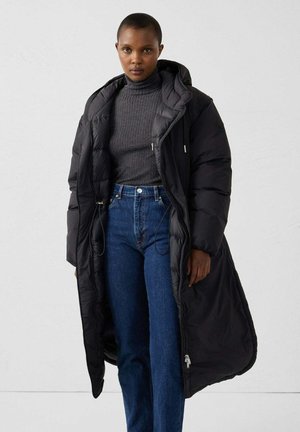 Lange zwarte gewatteerde jas met capuchon, voorzien van een ritssluiting, elastische trekkoorden en een getextureerde donkergrijze trui eronder. Blauwe jeans met hoge taille.