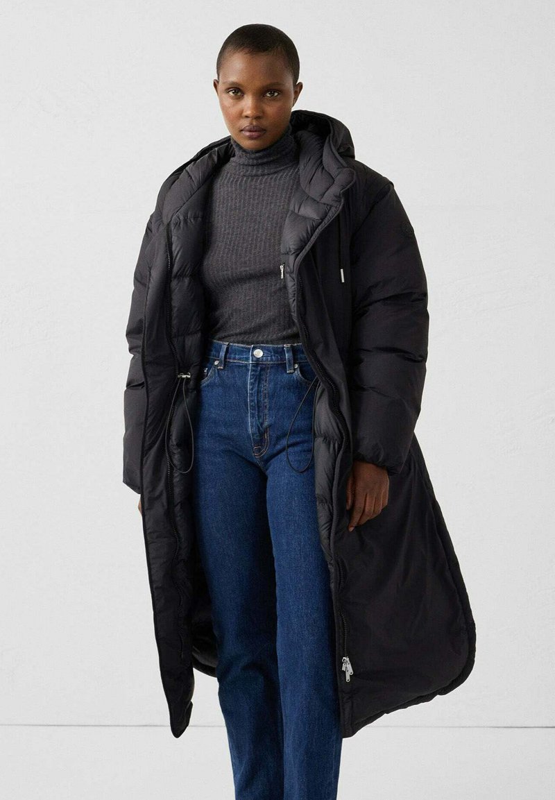 Langer schwarzer Steppmantel mit Kapuze, ausgestattet mit einem Reißverschluss, elastischen Kordelzügen und einem strukturierten dunkelgrauen Pullover darunter. Blaue High-Waist-Jeans.