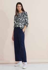 Blusa a righe bianco e nero con maniche arrotolate, abbinata a pantaloni larghi blu navy e sneakers bianche, su uno sfondo chiaro.