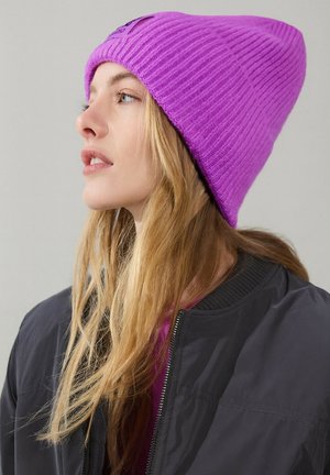 Fioletowa prążkowana dzianinowa czapka z ściągaczem, noszona z ciemną kurtką bomber z zamkiem błyskawicznym i prążkowanymi mankietami.