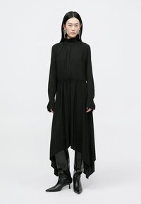 Vestido preto assimétrico feito de tecido fluido, apresentando um corpete franzido, mangas longas e uma barra distintivamente ampla. Combinado com botas pretas.