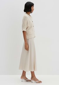 Un cardigan en tricot beige à manches courtes avec un col rond et trois boutons, associé à une jupe midi fluide de couleur beige clair et des sandales à talons argentés.