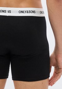 Nahaufnahme einer Person, die schwarze Boxershorts mit einem weißen Bund trägt, auf dem der Schriftzug "ONLY&SONS" zu sehen ist.