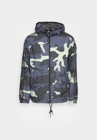 Giacca a vento con cappuccio in camouflage, blu marino e verde chiaro, realizzata in materiale leggero. Presenta una chiusura con zip frontale, orlo con cordino e tre strisce nere sulle maniche.