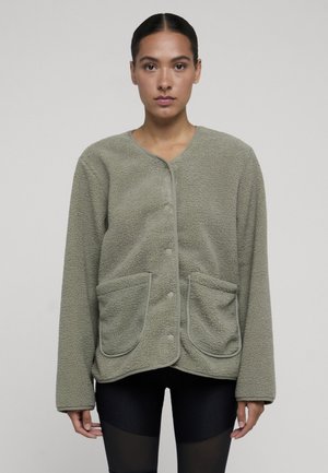 Femme portant une veste polaire vert clair avec des boutons-pression et de grandes poches avant, debout devant un fond uni.