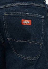 Τα σκούρα τζιν παντελόνια διαθέτουν μια πίσω τσέπη με κόκκινο λογότυπο Dickies, κίτρινη ραφή σε αντίθ εση και ελαφρώς υφασμένη όψη.