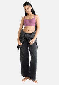 Ružový čipkovaný bralet s vlnkovaným lemom, v kombinácii s voľnými čiernymi džínsami a šedým sweatshirdom previazaným okolo pása. Nahé nohy, póza v stoji.