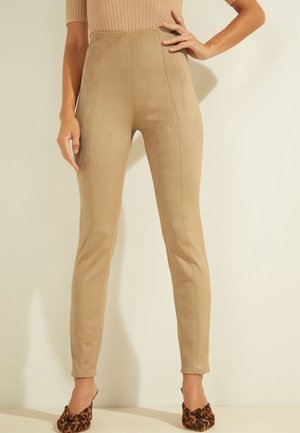 Vrouw draagt hoge taille leggings van beige suède, gecombineerd met leopardenprint mules met hak, staand tegen een lichte achtergrond.