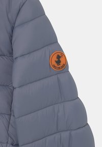 Šedá hřejivá bunda s prošívaným designem, hladkým povrchem a oranžovým kulatým štítkem s logem kachny na levém rukávu.