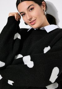 Schwarzer Pullover mit weißen Wolkenmustern, oversized Schnitt, gerippte Bündchen, weicher Stoff, über einem weißen Hemd mit Kragen getragen.