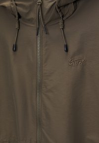 Chaqueta ligera color verde oliva con capucha, cremallera frontal, cordones elásticos y logo de la marca bordado en el pecho. Textura suave.