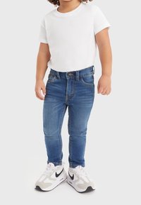 Vaqueros de mezclilla azul con corte ajustado, que cuentan con cinco bolsillos, detalles de costura y cierre de botón. Combinados con una camiseta blanca básica y zapatillas deportivas.