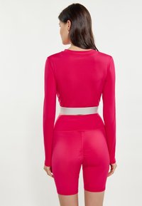 Top ajustat cu mâneci lungi și pantaloni scurți de bicicletă din material roz strălucitor și neted. Are o talie lată albă și un design fără cusături.