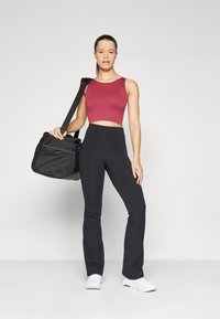 Rød atletisk crop top med tykke stropper, kombinert med høytlivede svarte flared leggings. Modellen holder en svart treningsbag. Hvite sneakers.