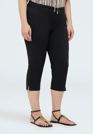 Persoon draagt zwarte cropped broeken met kleine zijslitsen en zwarte sandalen met gouden versieringen, staand tegen een effen achtergrond.