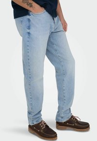 Man draagt lichtblauwe spijkerbroek, donkerbruine leren veterschoenen en een marineblauw overhemd met een zichtbare tatoeage op de onderarm, staand met één hand in de zak.