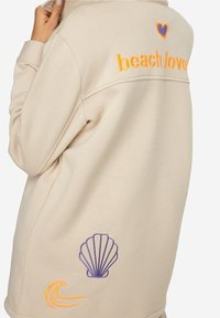 Sweat-shirt beige avec texte brodé orange "beach love", un cœur violet, un coquillage et un motif de vague au dos. Texture lisse.