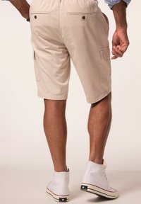 Beige cargo shorts har en strækbar talje, to baglommer med knapper og sidelommer; parret med hvide høje sko.