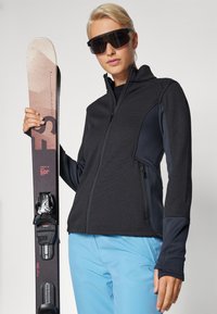 Crna fleece jakna s teksturiranim završetkom, plave skijaške hlače i skija u ruci. Velike naočale s tintom za sunce upotpunjuju sportski izgled.