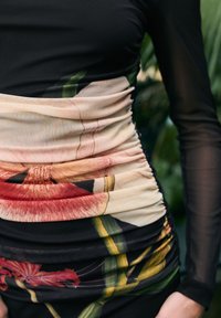 Czarna sukienka z kwiatowym nadrukiem w talii, z marszczeniem i przezroczystymi długimi rękawami. Tkanina ma gładką strukturę.