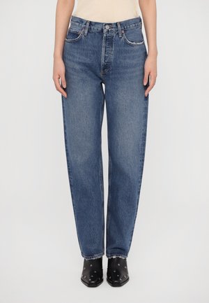 Vue de face d'une personne portant un jean bleu taille haute droit avec des ourlets effilochés et des bottines noires cloutées sur fond blanc.