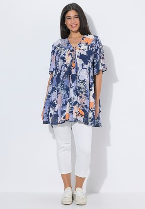 Kobieta uśmiechająca się, ubrana w luźną, niebieską tunikę w kwiaty, białe spodnie do połowy łydki i białe trampki, stojąca na tle jednolitego białego tła.