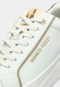 Λευκό δερμάτινο αθλητικό παπούτσι με χρυσές λεπτομέρειες, απαλή υφή, κρεμ κορδόνια και λογότυπο. Διαθέτει χρυσό περίγραμμα κατά μήκος της άκρης και διάτρητες λεπτομέρειες.