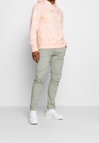 Peach tie-dye hoodie with front pocket, paired with light green slim-fit pants. White sneakers with green accent on heel.