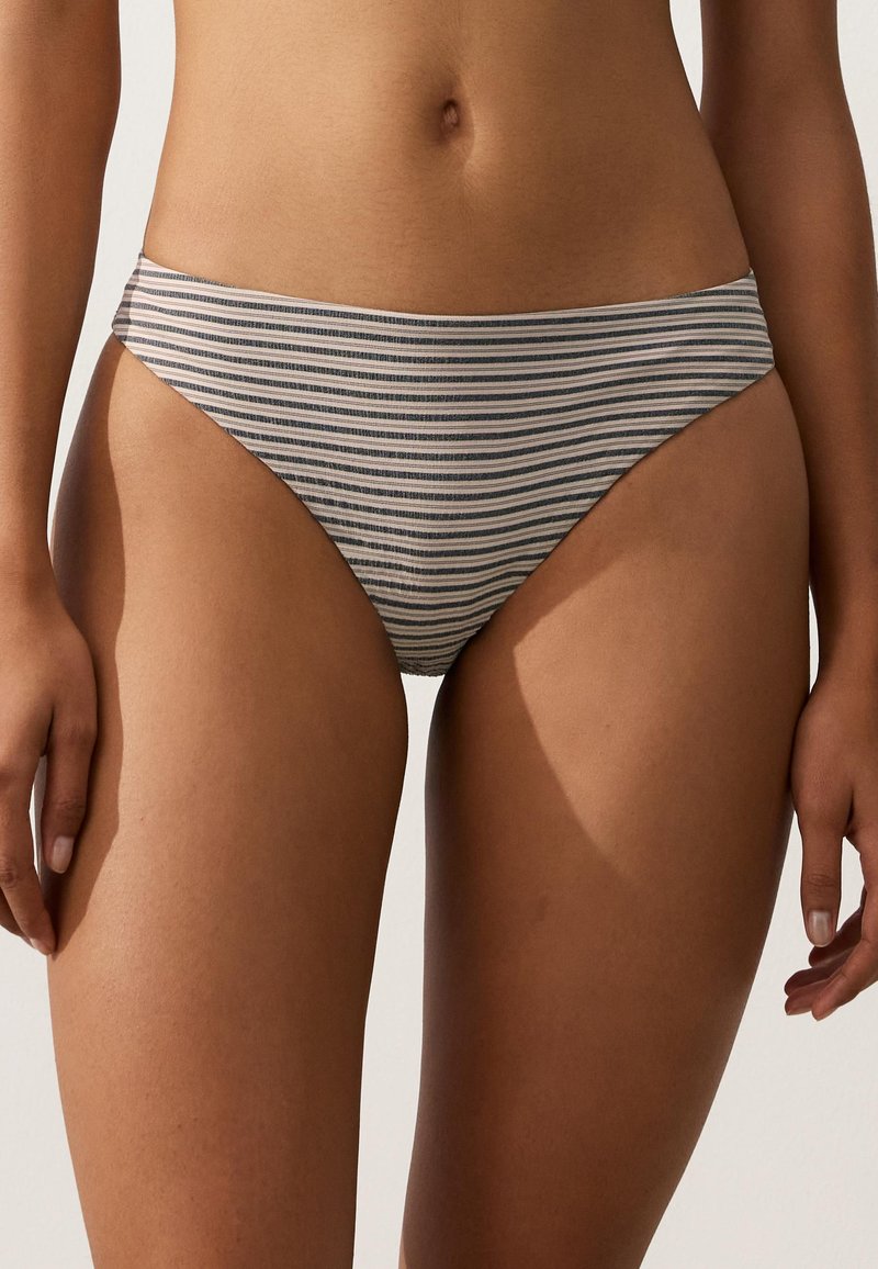 Femme portant un bas de bikini rayé gris et blanc, debout, les mains relâchées le long du corps sur un fond uni.