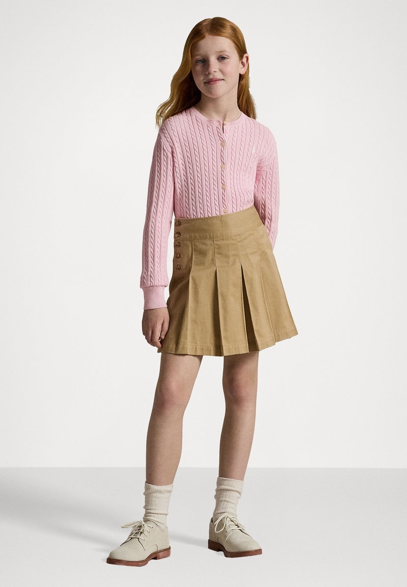 Cárdigan de punto rosa, falda plisada beige con botones laterales, calcetines blancos y zapatos claros. Diseño simple, tejido texturizado y colores neutros.