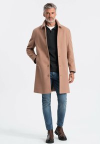 Abrigo de lana beige con cuello y botones en el frente, combinado con un suéter negro y jeans azules. La modelo está de pie con las manos en los bolsillos.