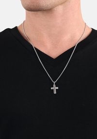 Ciondolo a forma di croce in argento appeso a una sottile catena, indossato su una maglietta nera a scollo a V. Il ciondolo presenta un design semplice e liscio con una finitura lucida.