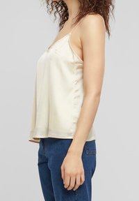 Light cream satin camisole featuring thin straps, a v-neckline, and a loose fit, paired with dark blue denim jeans.