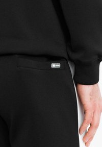 Primo piano della mano di una persona vicino alla tasca posteriore di pantaloni neri, che mostra una piccola patch rettangolare con il logo sul bordo della tasca.