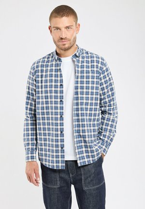 Homme portant une chemise boutonnée à carreaux bleus et blancs sur un t-shirt blanc avec un pantalon en denim foncé sur un fond uni.