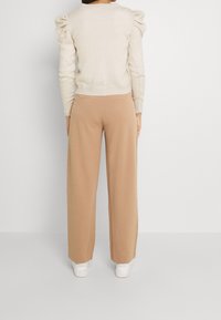 Beżowy sweter z dzianiny z bufiastymi ramionami, zestawiony z jasnobrązowymi spodniami o szerokich nogawkach. Ubranie dopełniają białe sneakersy oraz dopasowany krój.