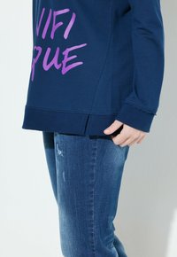 Felpa blu navy con testo grafico viola, abbinata a jeans blu strappati. La felpa ha un orlo dritto e fessure laterali.
