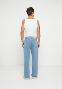 Hellblaue weite Jeans mit klassischem Fünf-Taschen-Design, kombiniert mit einem weißen Tanktop und beigen Schuhen vor einem neutralen Hintergrund.