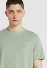 Maglietta a maniche corte verde chiaro realizzata in tessuto morbido. Presenta un colletto rotondo e un piccolo logo giallo sull'area sinistra del petto.