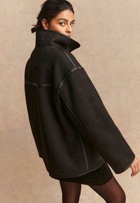 Czarna oversize kurtka z wysokim kołnierzem, wykonana z miękkiego materiału. Posiada kontrastowe skórzane akcenty wzdłuż szwów.
