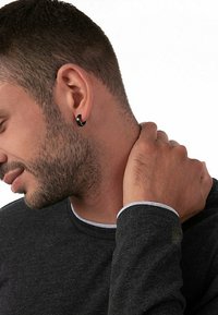 Maglietta nera a maglia con rifiniture grigie chiare, che presenta un uomo con un piccolo orecchino a cerchio nero che si tocca il collo. Texture della pelle liscia visibile.