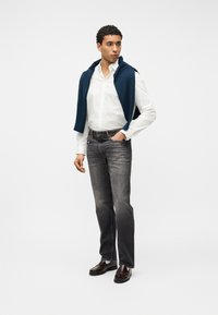 Νεαρός άνδρας με λευκό πουκάμισο, τζιν με σκουρόχρωμο ξέβαμμα, καφέ παπούτσια, με ένα μπλε πουλόβερ περασμένο στους ώμους, στέκεται μπροστά σε ομοιόμορφο φόντο.