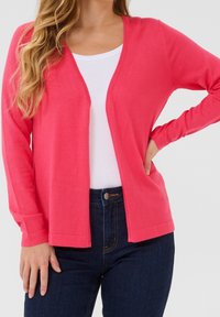 Lichtgewicht koraalkleurig vest gemaakt van een gestructureerde stof, met een V-hals, open voorkant en lange mouwen, gecombineerd met een wit topje en donkere jeans.