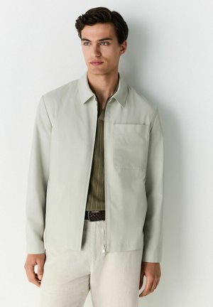 Chaqueta gris claro de tela texturizada, con cuello, cremallera frontal y bolsillo en el pecho izquierdo. Lleva encima una camisa verde.