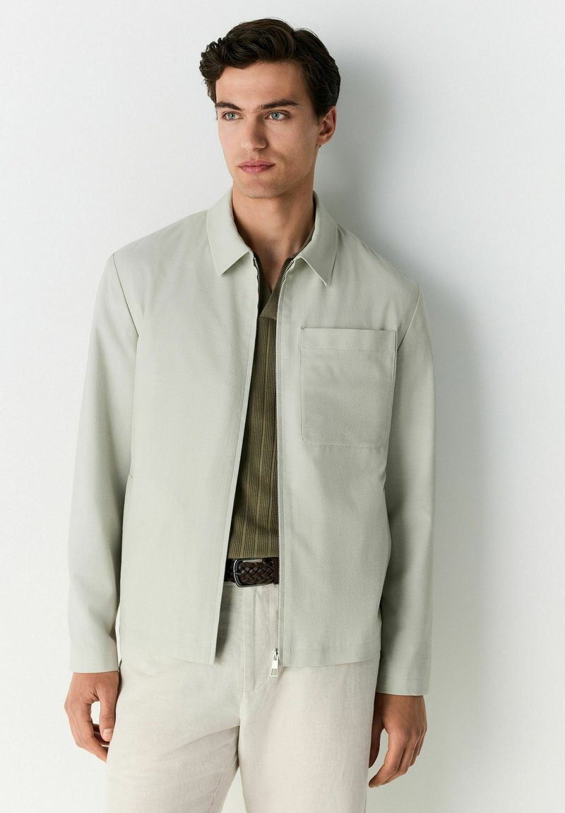 Chaqueta gris claro de tela texturizada, con cuello, cremallera frontal y bolsillo en el pecho izquierdo. Lleva encima una camisa verde.