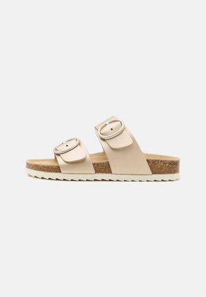 Beige slip-on sandal med to justerbare stropper med ovale sølvspænder, kork-fodsål og hvid rillesål i gummi.