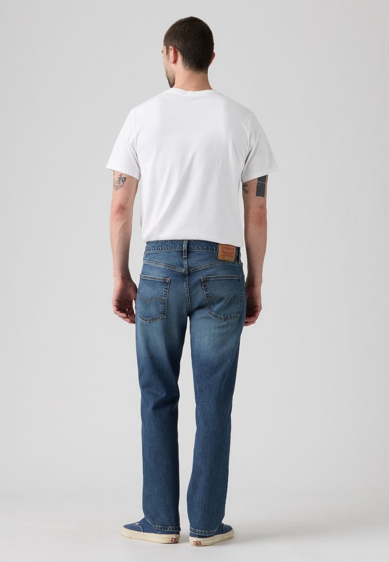 Man standing facing away wearing a plain white t-shirt, blue jeans, and blue sneakers on a plain light grey background.
