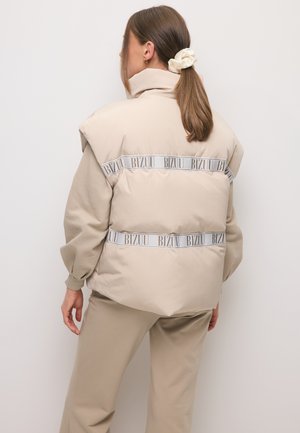 Vrouw met bruin haar in een paardenstaart, gekleed in een beige gevoerd vest met "BIZUU" tape details, gedragen over een bijpassende beige top met lange mouwen en broek.