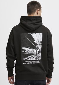 Hoodie noir avec un grand motif abstrait blanc au dos, représentant une scène urbaine floue et du texte. Le tissu semble doux et épais.